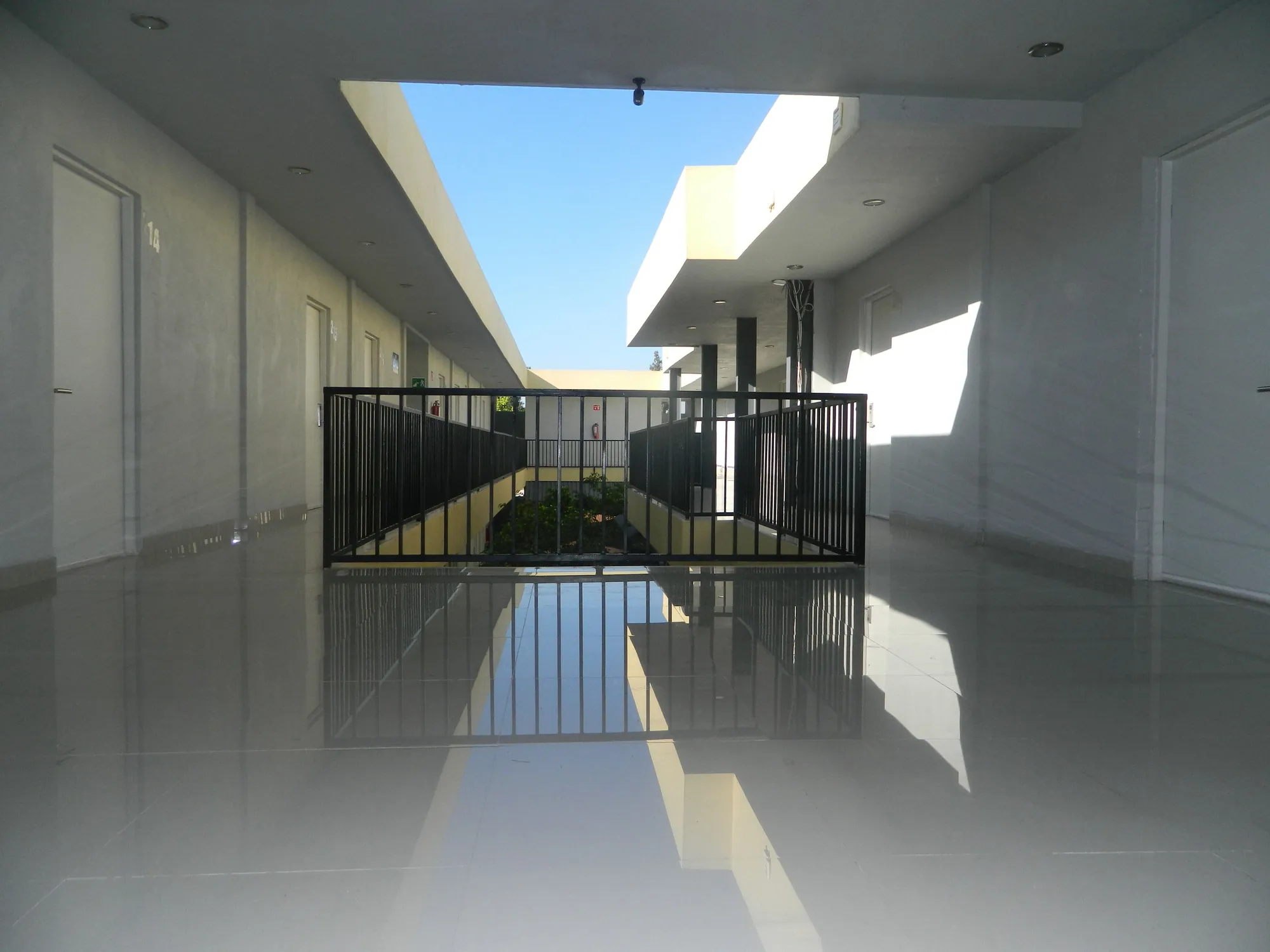 Pasillo de piso brillante que da al patio central, con barandal negro y el cielo azul arriba